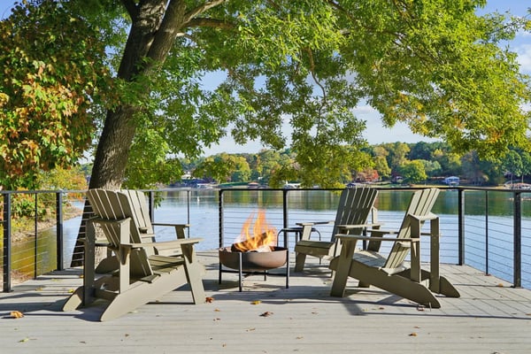 Waterfront deck with Adirondack chairs surrounding a fire pit, large tree overhead, river view with forested shoreline