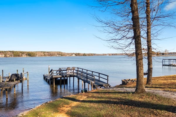 Wooden dock and boat launch on calm lake with bare trees and distant shoreline homes under clear blue sky