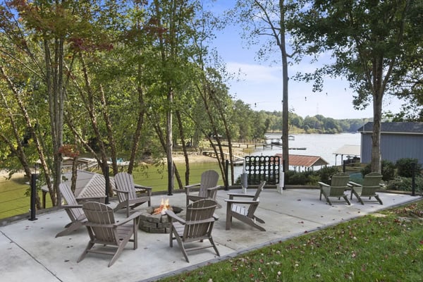 Waterfront patio with Adirondack chairs around fire pit, lake view, tall trees, and dock in background