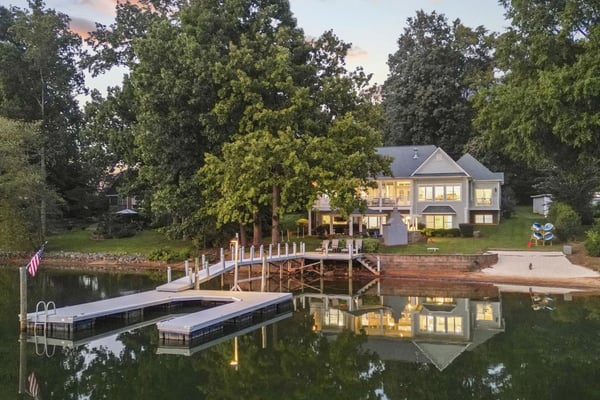 Waterfront home at dusk with lit windows, wooden dock, floating platforms, and mature trees surrounding the property.