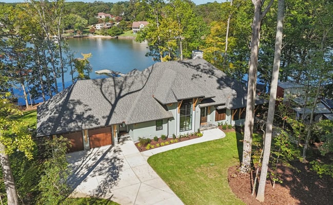 Modern lakefront home with gray roof and white exterior surrounded by tall trees, overlooking calm blue water with residential properties visible across the